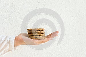 Young caucasian woman holds in stretched hand stack of chocolate chip cookies with oats bran nuts on white wall. Holiday baking
