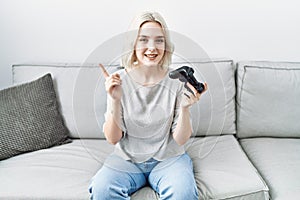 Young caucasian woman at home playing video game holding controller smiling happy pointing with hand and finger to the side
