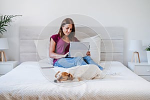 Young caucasian woman at home working on laptop with cute jack russell dog resting on bed. Home office, Pets, love and relax.