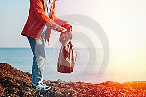 A young Caucasian woman in a jacket collects garbage in a plastic bag on the beach. In the background, the horizon of the sea and