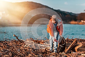 A young Caucasian woman in a jacket collects garbage in a plastic bag on the beach. In the background, the sea and mountains.