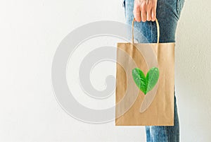 Young caucasian woman in jeans holds in hand empty blank mock up brown craft paper bag on white wall background. Heart logo leaves