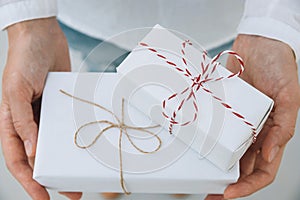 Young caucasian woman in jeans shirt holds in hands stacked gift boxes wrapped in white paper tied with striped twine. Christmas