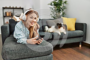 Young caucasian woman listening to music lying on sofa with dog at home