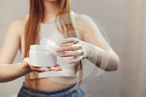 Young caucasian woman with long flowing hair is opening unbranded white jar with cosmetic cream