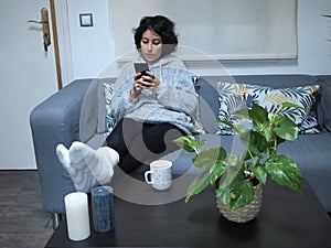 Young caucasian woman looking at her phone with her feet on the table, in pajamas, sitting on the sofa with a cup of coffee