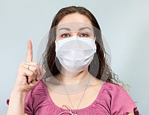 Young Caucasian woman in medical face protection mask gesturing with forefinger on blue background, looking at camera