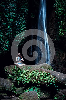 Young Caucasian woman meditating, practicing yoga at waterfall. Hands in namaste mudra. Leke Leke waterfall, Bali, Indonesia