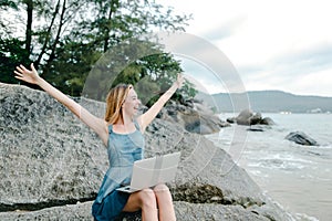 Young caucasian woman with outstretched hands working with laptop on sea shore and sitting on stone.