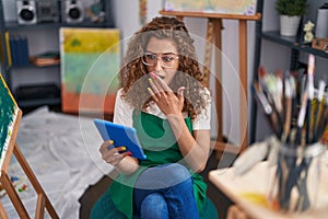 Young caucasian woman painter sitting at art studio doing video call with tablet covering mouth with hand, shocked and afraid for