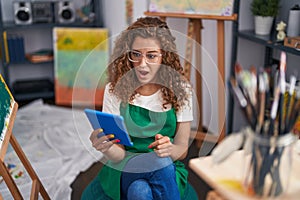 Young caucasian woman painter sitting at art studio doing video call with tablet scared and amazed with open mouth for surprise,