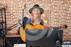 Young caucasian woman playing classic guitar at music studio pointing thumb up to the side smiling happy with open mouth