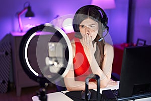 Young caucasian woman playing video games recording with smartphone covering mouth with hand, shocked and afraid for mistake