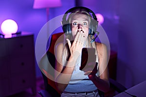 Young caucasian woman playing video games with smartphone covering mouth with hand, shocked and afraid for mistake