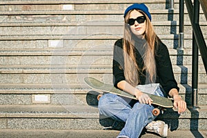 Young caucasian woman posing on street with skateboard in hands. Teenager girl in blue jeans extreme sports in an urban