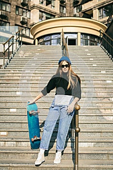 Young caucasian woman posing on street with skateboard in hands. Teenager girl in blue jeans extreme sports in an urban