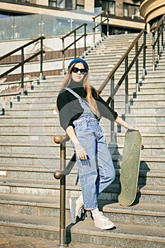 Young caucasian woman posing on street with skateboard in hands. Teenager girl in blue jeans extreme sports in an urban