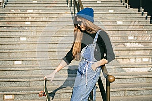 Young caucasian woman posing on street with skateboard in hands. Teenager girl in blue jeans extreme sports in an urban