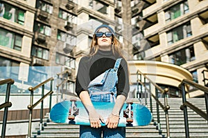 Young caucasian woman posing on street with skateboard in hands. Teenager girl in blue jeans extreme sports in an urban