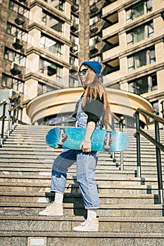 Young caucasian woman posing on street with skateboard in hands. Teenager girl in blue jeans extreme sports in an urban