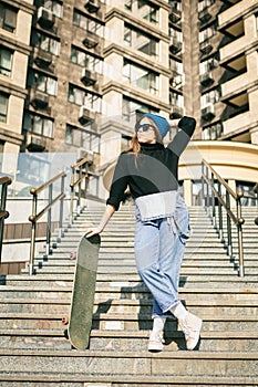 Young caucasian woman posing on street with skateboard in hands. Teenager girl in blue jeans extreme sports in an urban