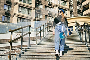 Young caucasian woman posing on street with skateboard in hands. Teenager girl in blue jeans extreme sports in an urban