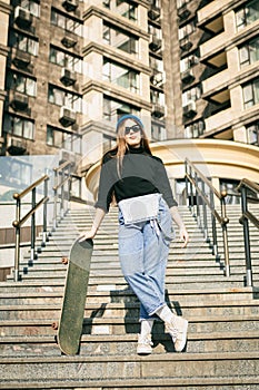 Young caucasian woman posing on street with skateboard in hands. Teenager girl in blue jeans extreme sports in an urban