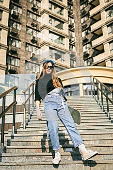 Young caucasian woman posing on street with skateboard in hands. Teenager girl in blue jeans extreme sports in an urban