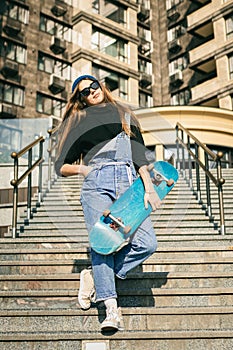 Young caucasian woman posing on street with skateboard in hands. Teenager girl in blue jeans extreme sports in an urban