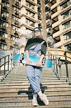 Young caucasian woman posing on street with skateboard in hands. Teenager girl in blue jeans extreme sports in an urban