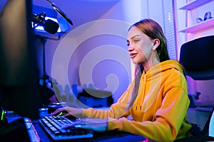 Young Caucasian woman professional gamer wearing yellow hoodie sits on a chair with a gaming table with pc, keyboard, monito?.