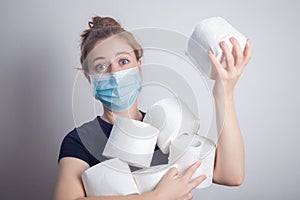 Young Caucasian woman in protective mask holding many toilet paper rolls. Panic hoarding, buying for the coronavirus quarantine