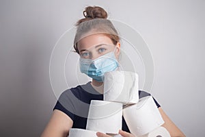 Young Caucasian woman in protective mask holding many toilet paper rolls. Panic hoarding, buying for the coronavirus quarantine