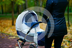 Young caucasian woman is pushing white baby carriage while she walking at autumn park with beautiful yellow leaves