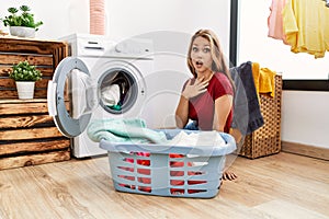 Young caucasian woman putting dirty laundry into washing machine scared and amazed with open mouth for surprise, disbelief face