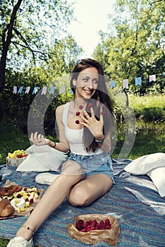 Young Caucasian woman with raspberries on her fingers smiling and posing on a picnic blanket