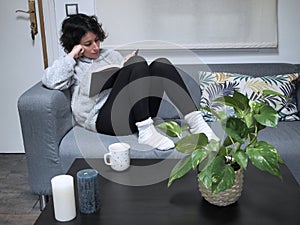 Young caucasian woman reading a book in pajamas and socks, sitting on the sofa with a cup of coffee