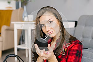 Young Caucasian woman in a red plaid shirt and skirt does her makeup, paints her lips with glitter at home, sitting on the couch