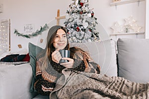 Young caucasian woman relaxing on a sofa at home and holding mug with hot drink in Christmas holidays