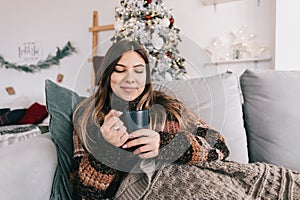 Young caucasian woman relaxing on a sofa at home and holding mug with hot drink in Christmas holidays