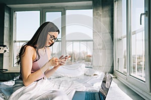 Young caucasian woman resting on a bed at home and using laptop computer and smartphone. Chatting with friends, shopping online