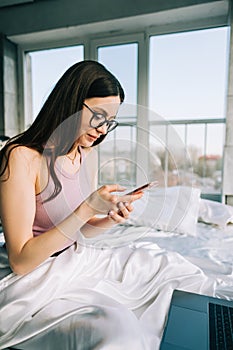 Young caucasian woman resting on a bed at home and using laptop computer and smartphone. Chatting with friends, shopping online