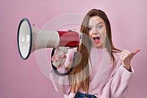 Young caucasian woman shouting through megaphone pointing thumb up to the side smiling happy with open mouth