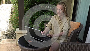 Young Caucasian woman sitting in armchair, typing on laptop and drinking coffee from mug while working in home garden