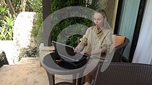 Young Caucasian woman sitting in armchair, typing on laptop and drinking coffee from mug while working in home garden