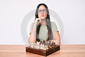 Young caucasian woman sitting at the desk playing chess serious face thinking about question with hand on chin, thoughtful about