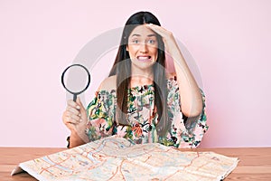 Young caucasian woman sitting at the desk searching on city map using magnifying glass stressed and frustrated with hand on head,