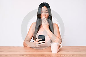 Young caucasian woman sitting at the desk using smartphone drinking coffee covering mouth with hand, shocked and afraid for