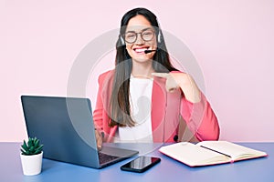 Young caucasian woman sitting at the desk wearing call center agent headset working using laptop pointing finger to one self