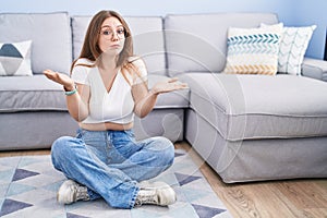 Young caucasian woman sitting on the floor at the living room clueless and confused expression with arms and hands raised